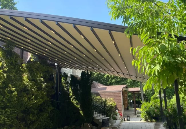 Pergolă retractabilă Arcodia instalată în curte cu vegetație, Brașov Romania Pergolă retractabilă Arcodia cu iluminare LED, instalată într-o grădină verde din Brașov, România.